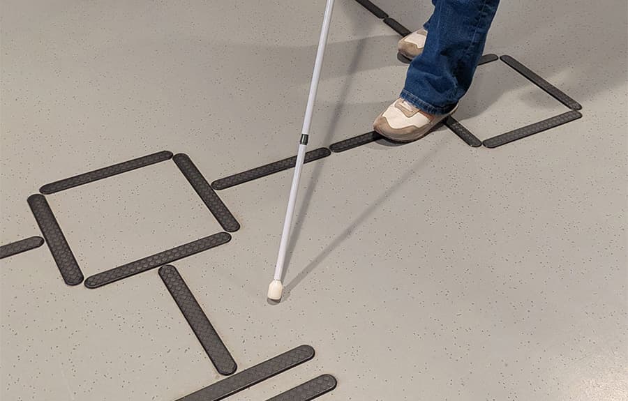 Bena på en person som står på et innendørs ledelinjesystem med vendefelt og oppmerksomhetsfelt. Personen har mobilitetsstokk.