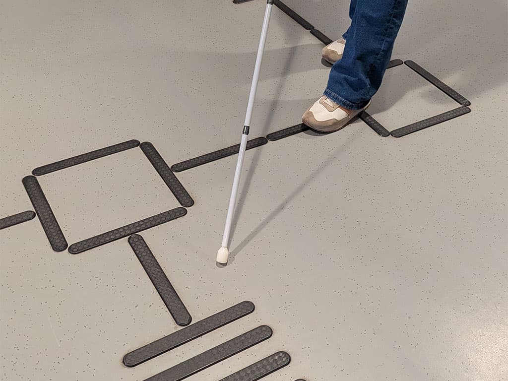 To ben og en mobilitistokk står på en ledelinje. Vendefelt og oppmersomhetsfelt melleom ledelinjene.