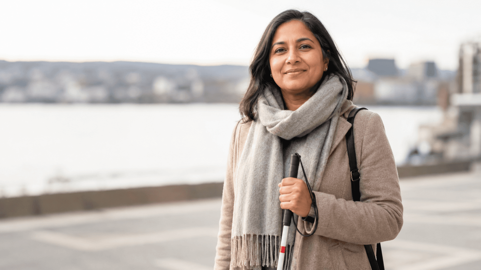 Smilende kvinne med mørkt hår holder en hvit stokk på en promenade ved vannet, med byen uskarpt i bakgrunnen.