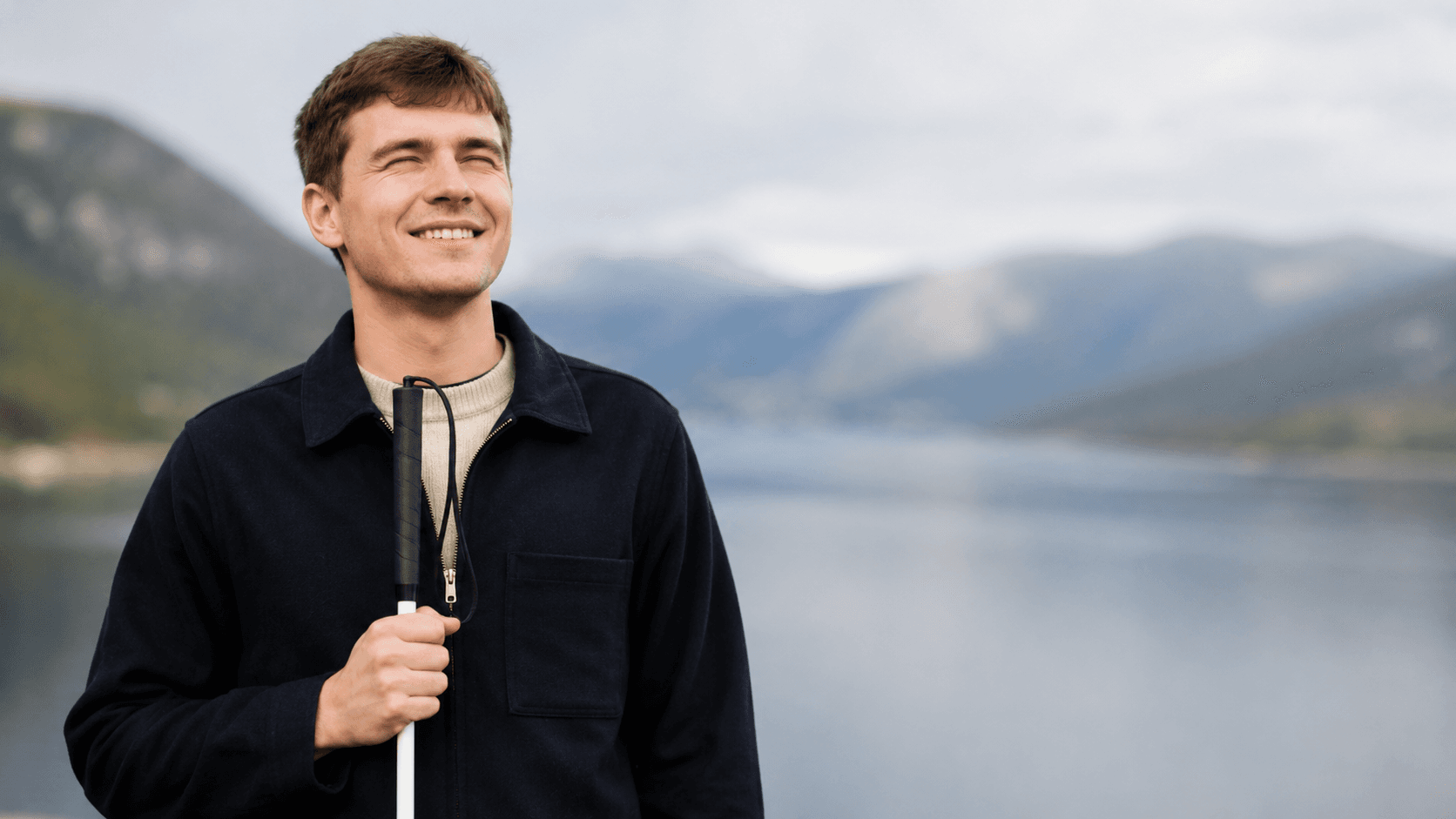 Ung mann med hvit stokk smiler utendørs foran et fjordlandskap med fjell og vann i bakgrunnen.