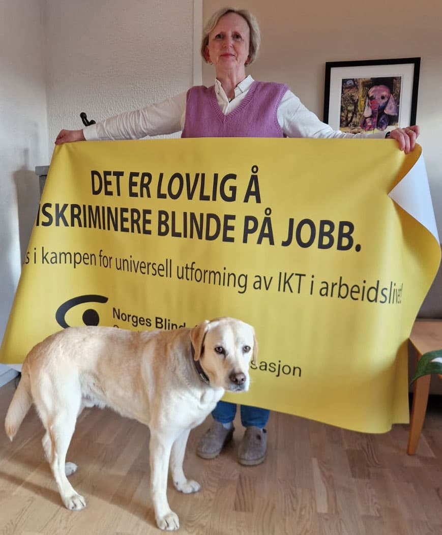 Birgit Risholt holder parolen, foran den står det en lys labrador. Tekst: DET ER LOVLIG Å DISKRIMINERE BLINDE PÅ JOBB. Støtt oss i kampen for universell utforming av IKT i arbeidslivet.