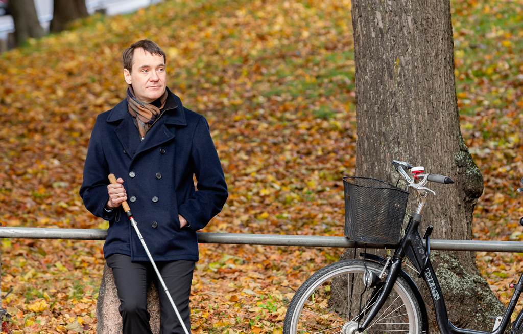 Per Inge Bjerknes i høstmiljø med hvit stokk. Han sitter på et gjerde, ved siden av et tre og en parkert sykkel.
