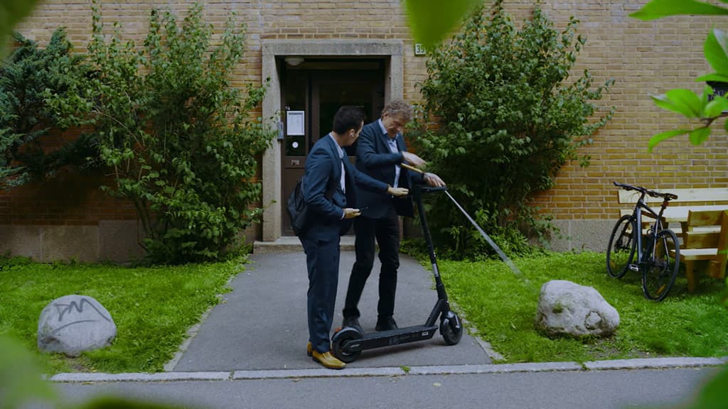 En mann med hvit stokk er i ferd med å ta fra en mann i dress en elsparkesykkel.