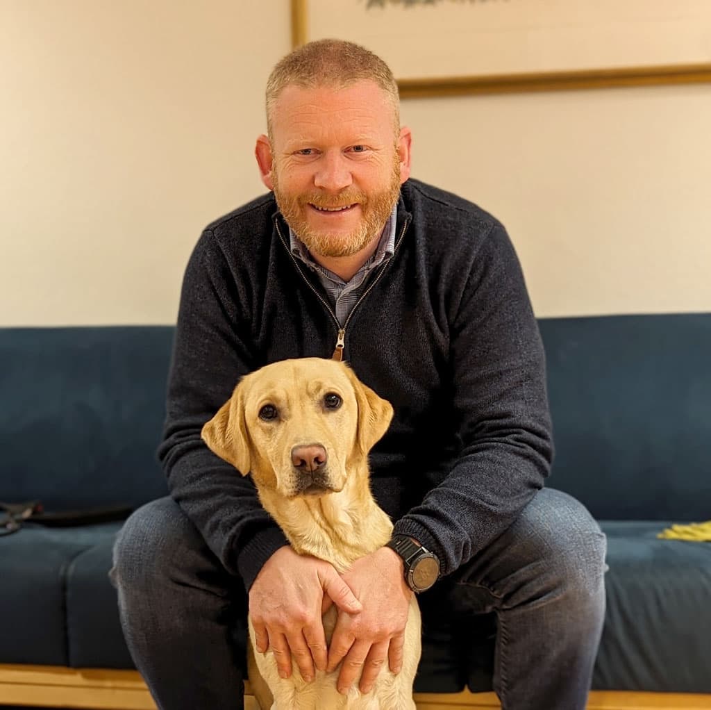 Tommy sitter i en sofa med en lys labrador på gulvet foran seg.