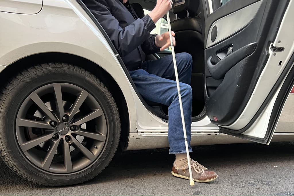 En person med hvit stokk er i ferd med å sette seg inn i en taxi.