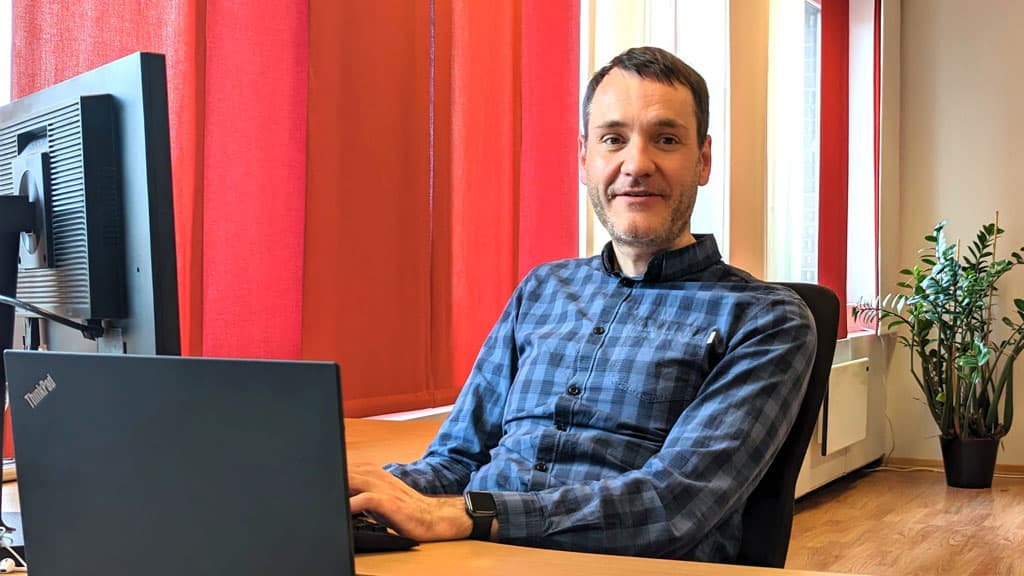 Per Inge Bjerknes bak skrivebordet ved PCen i rutetet skjorte. Det er røde gardiner og en stor, grønn plante i bakgrunnen