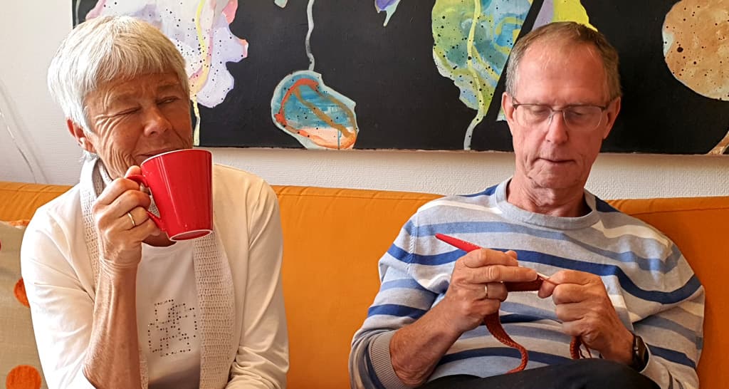 Torhild og Arne sitter i en sofa. Torhild drikker kaffe og Aren strikker.