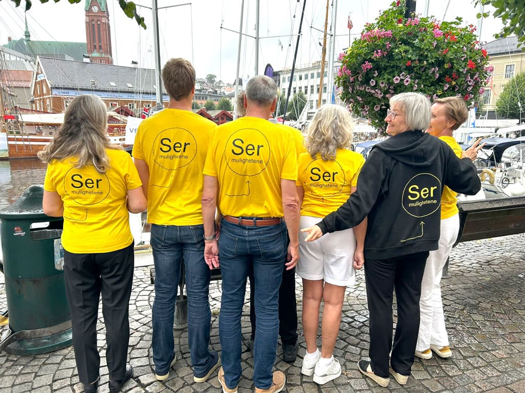 En gruppe mennesker med gule t-skjorter står med ryggen mot kamera i havnen i Arendal. På ryggene deres står det "Ser mulighetene"