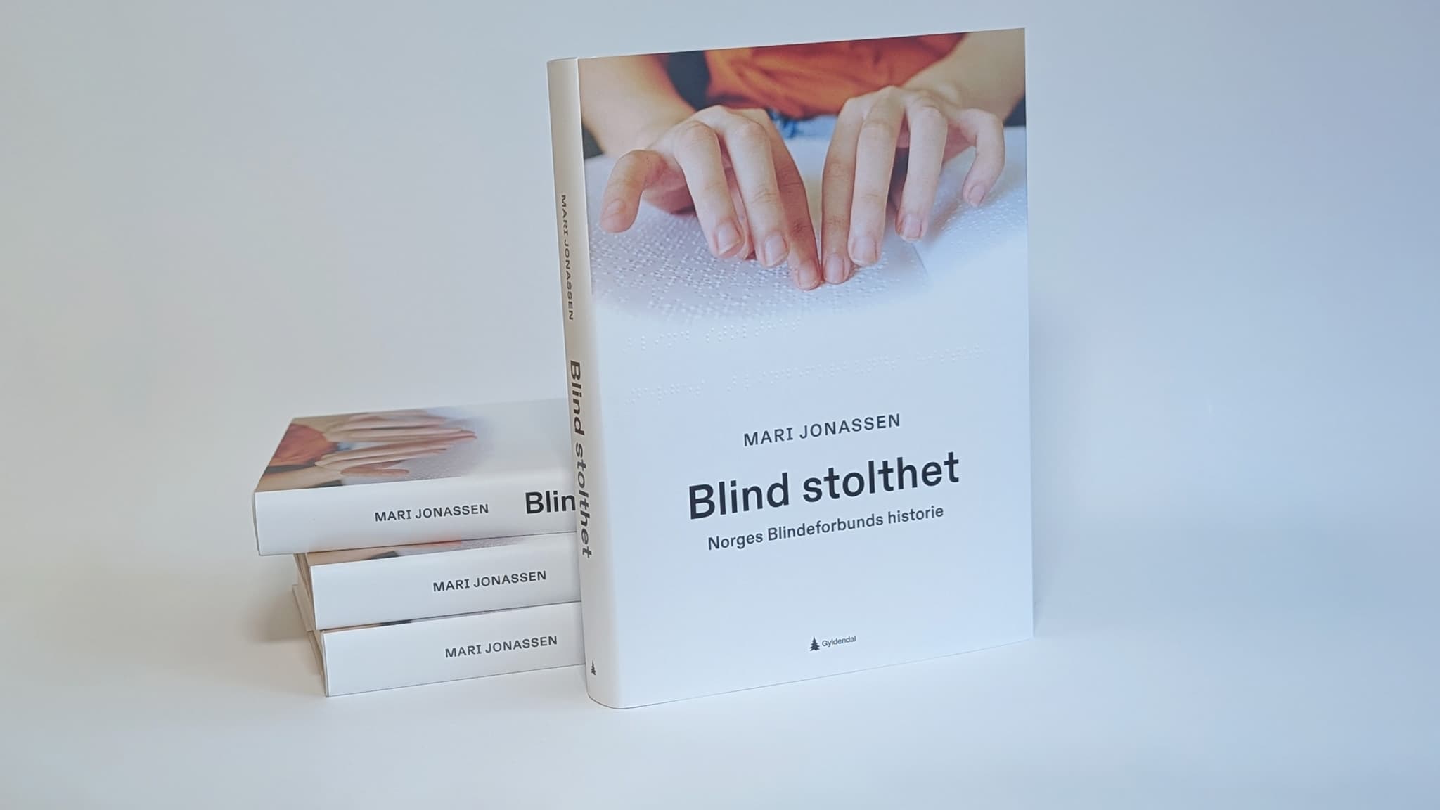 Bøker i stabel og forsiden av boka, hender som leser punktskrift, tekst "Blind stolthet", Mari Jonassen, Norges Blindeforbunds historie.